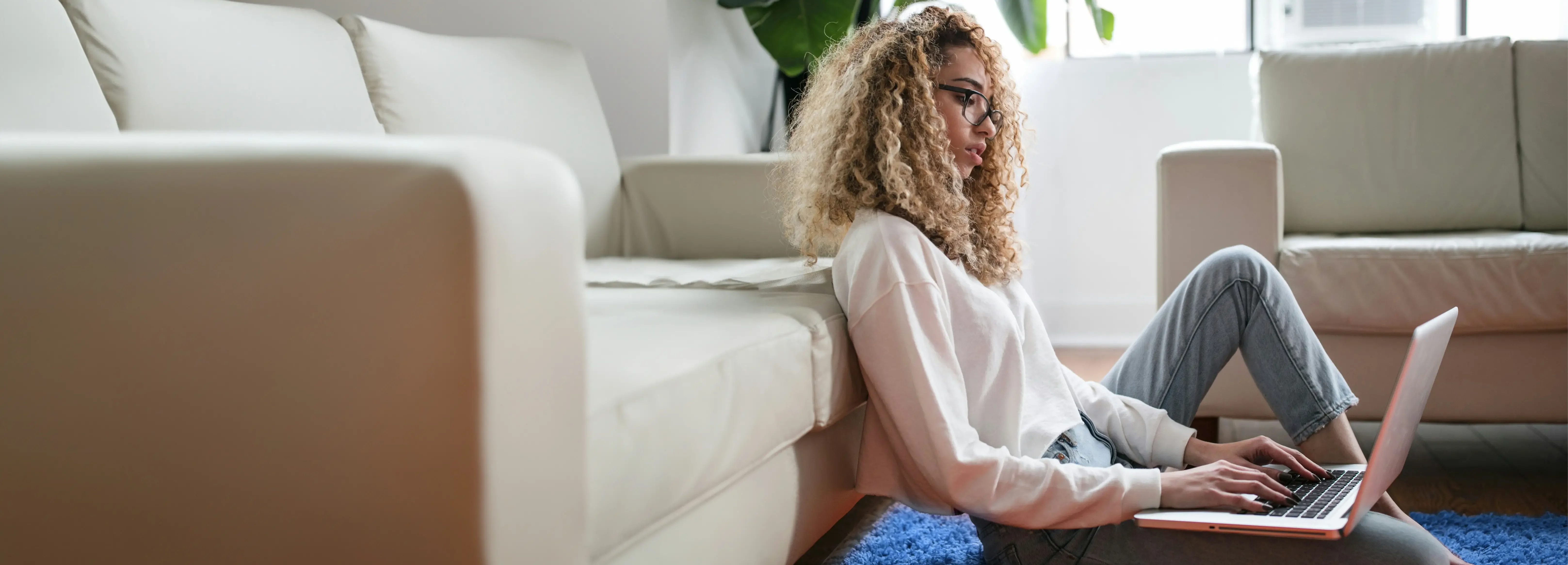 Junge Frau mit blonden Locken sitzt auf dem Teppich vor einem Sofa und arbeitet an einem Laptop.