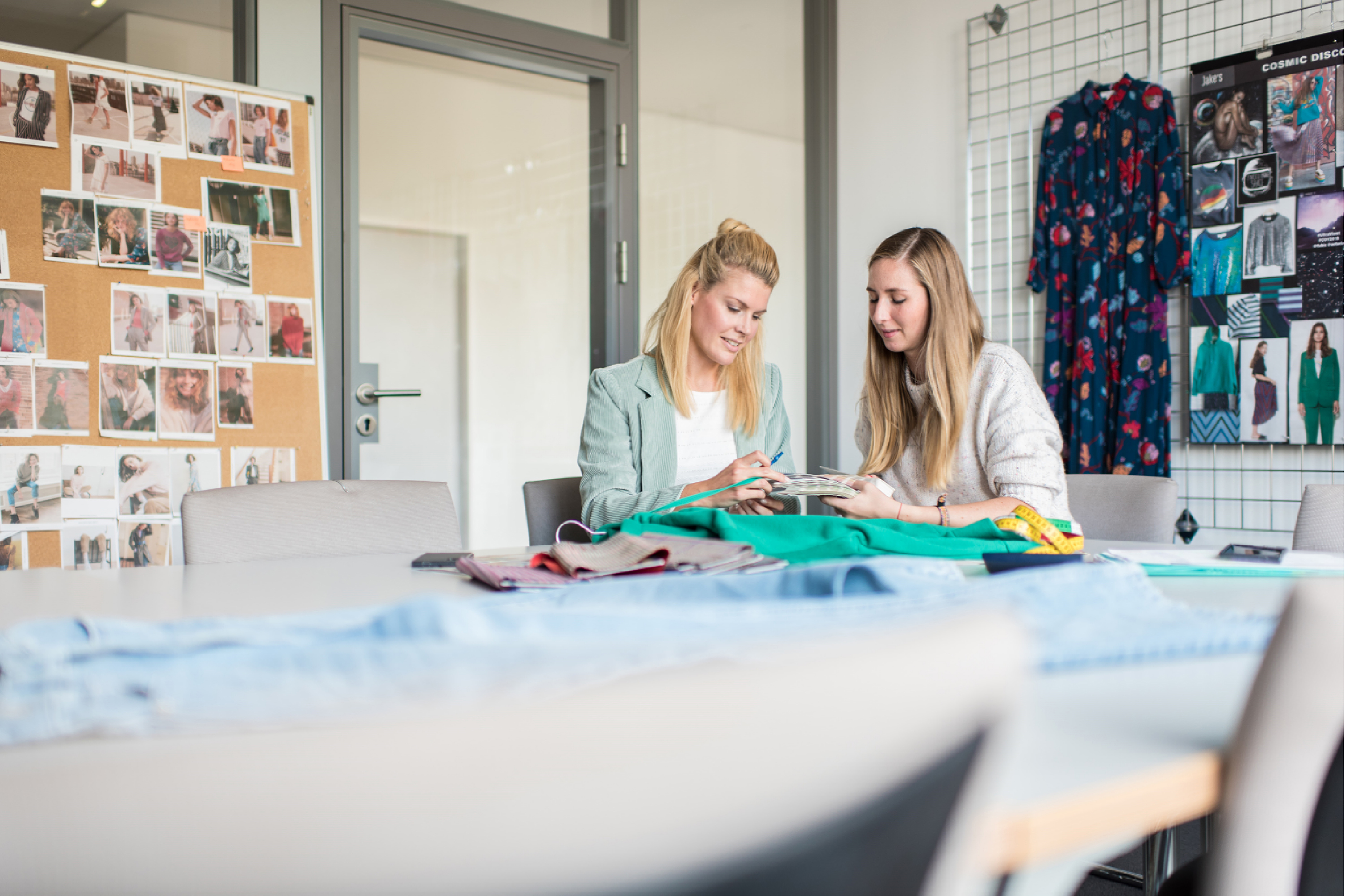 Zwei  Einkäuferinnen sitzen an einem Tisch und betrachten Stoffe und Ware. Kreatives Umfeld mit Mood Boards und Kleidungsstücken die im Hintergrund hängen.