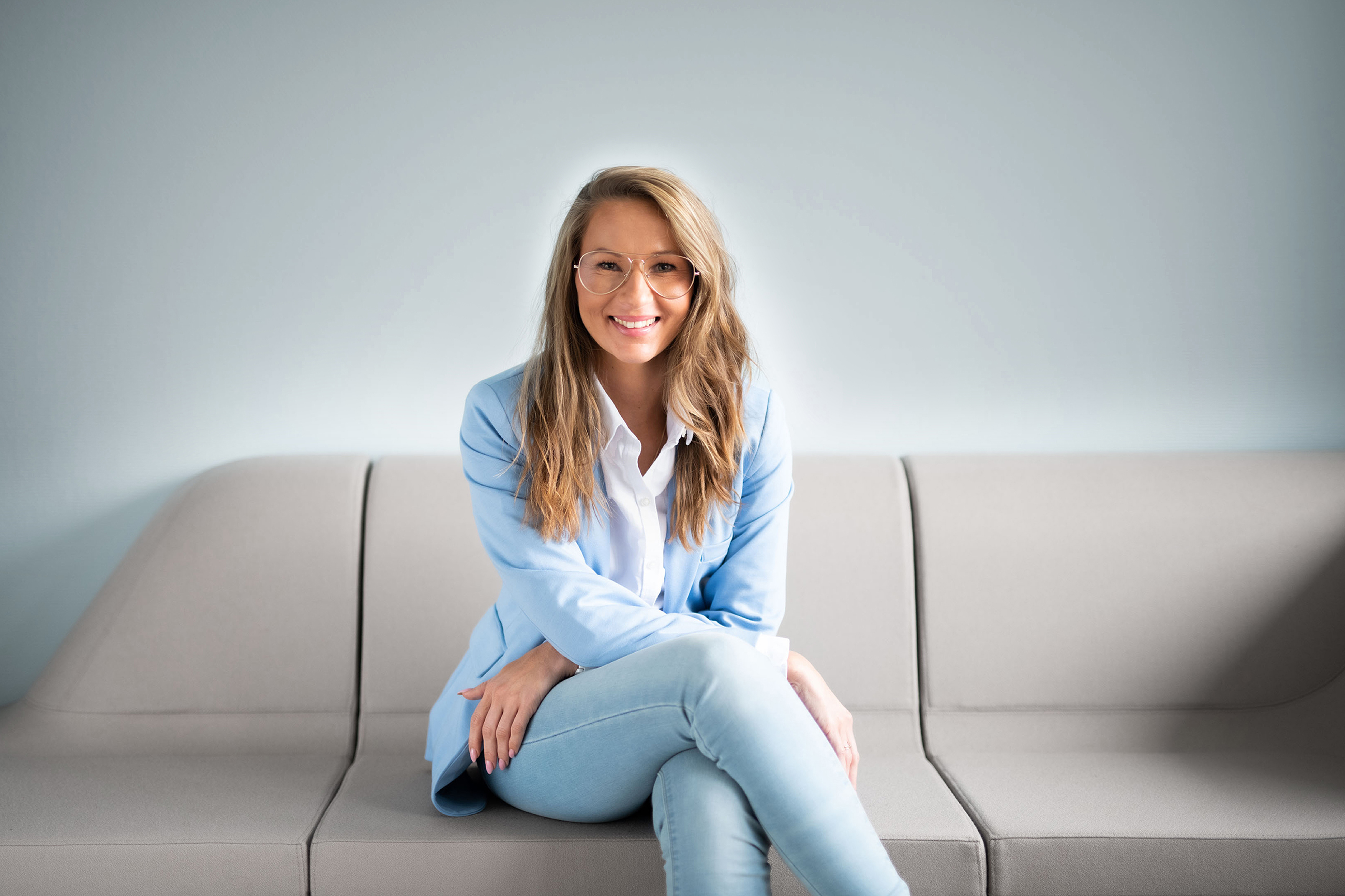 Junge Frau mit Brille sitzt in hellblau gekleidet auf einem modernen Sofa und blickt lächelnd in die Kamera. 
