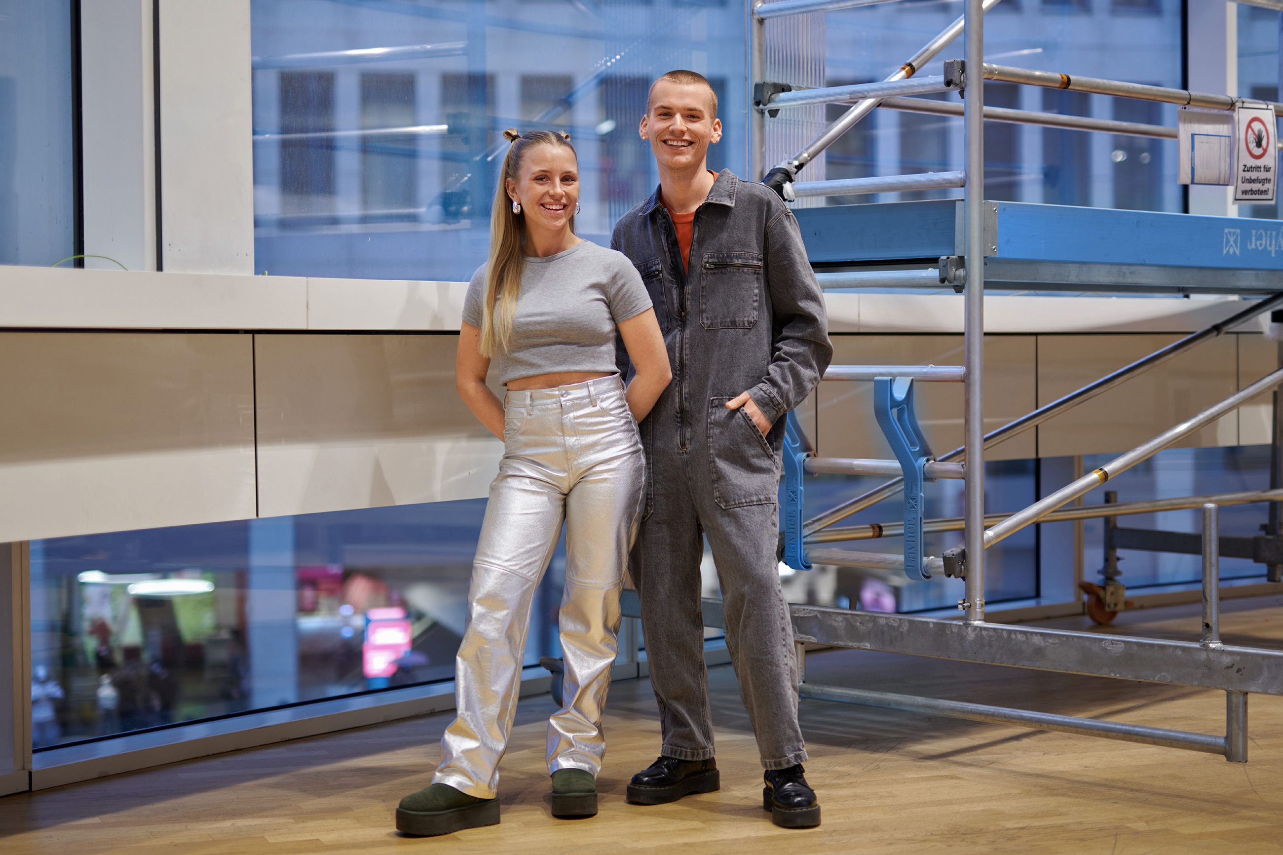 Zwei Gestalter-Azubis modern gekleidet in silber und Jeans Jumpsuit stehen vor einem Gerüst und lächeln in die Kamera