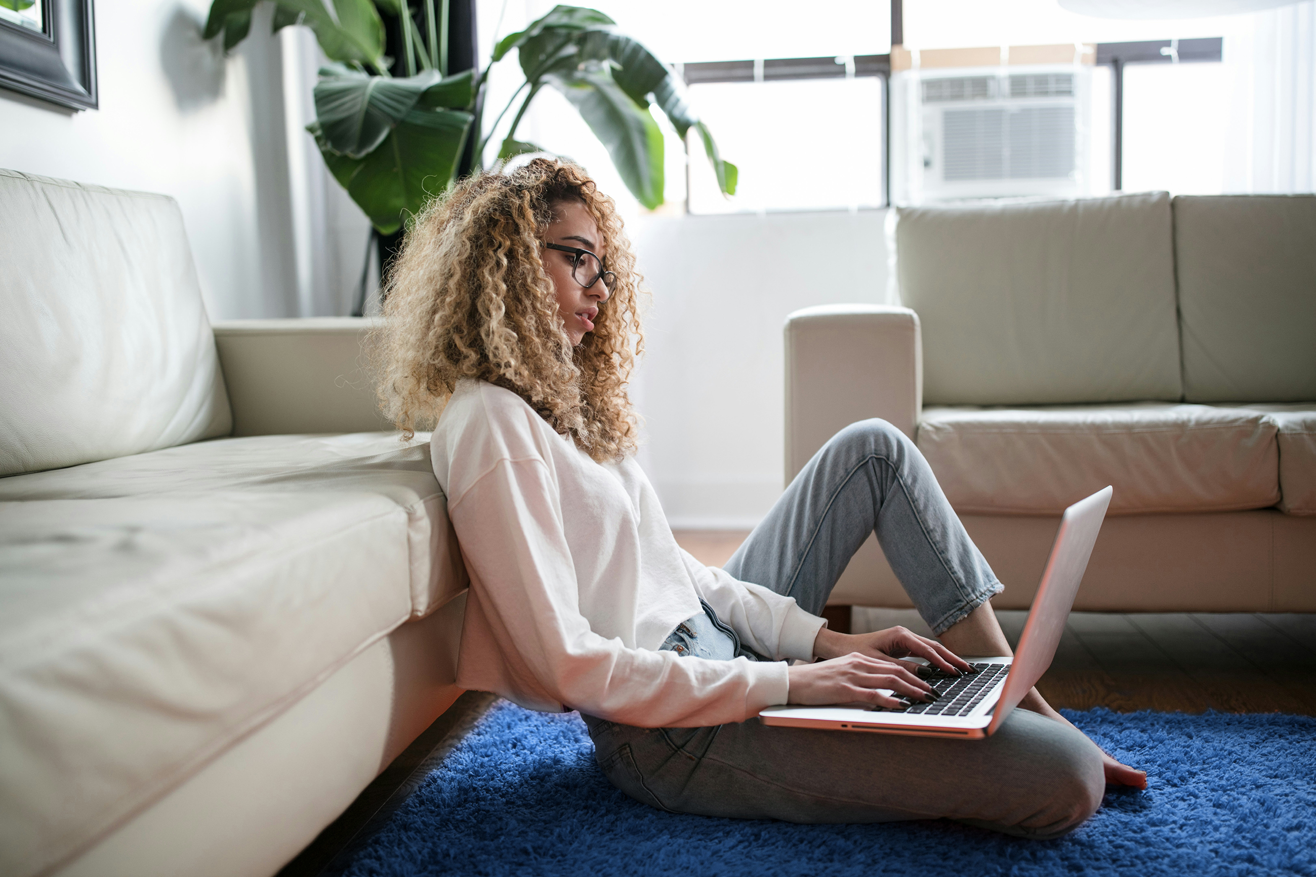 Frau mit langen blonden Locken sitzt auf dem Teppich in ihrer Wohnung und arbeitet an einem Laptop auf ihrem Schoß
