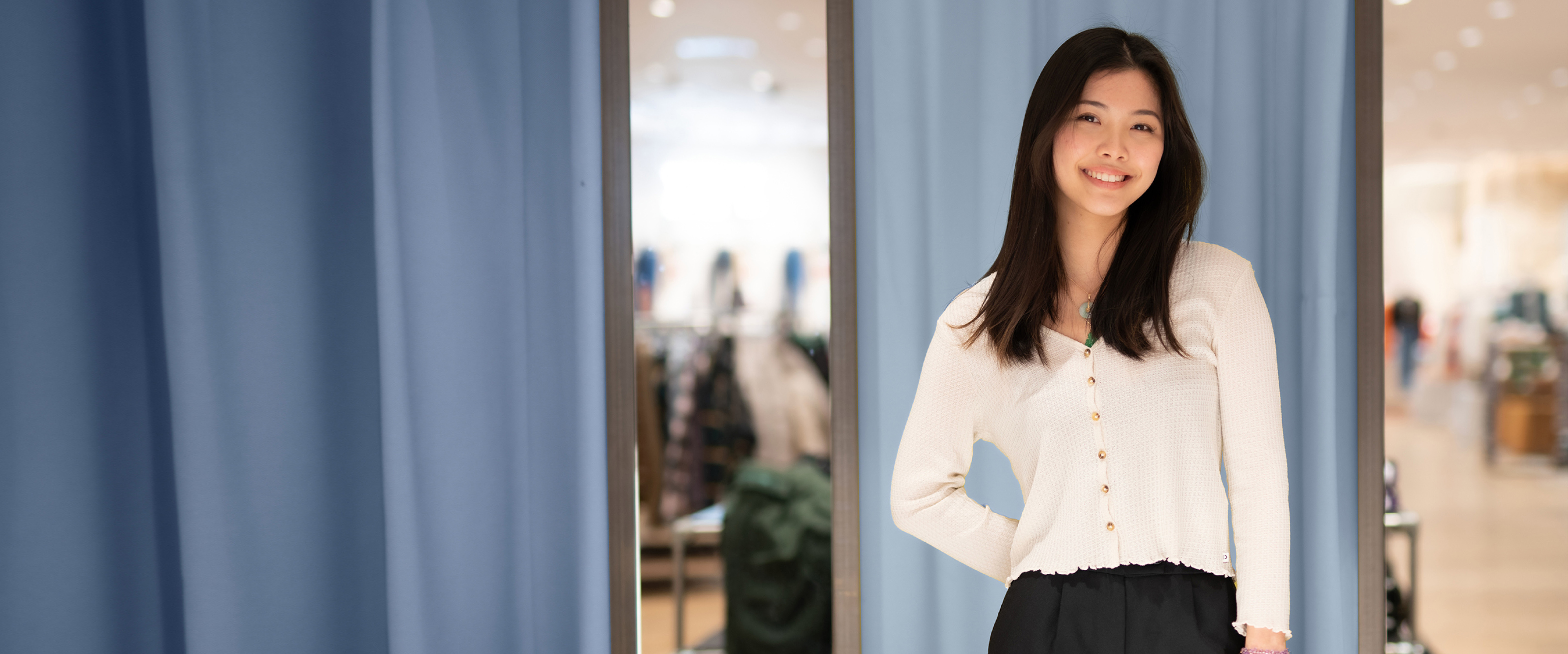 Junge Auszubildende mit schwarzen Haaren, asiatisch gelesen, modern gekleidet steht im Verkaufshaus vor Garderoben in Blautönen. 