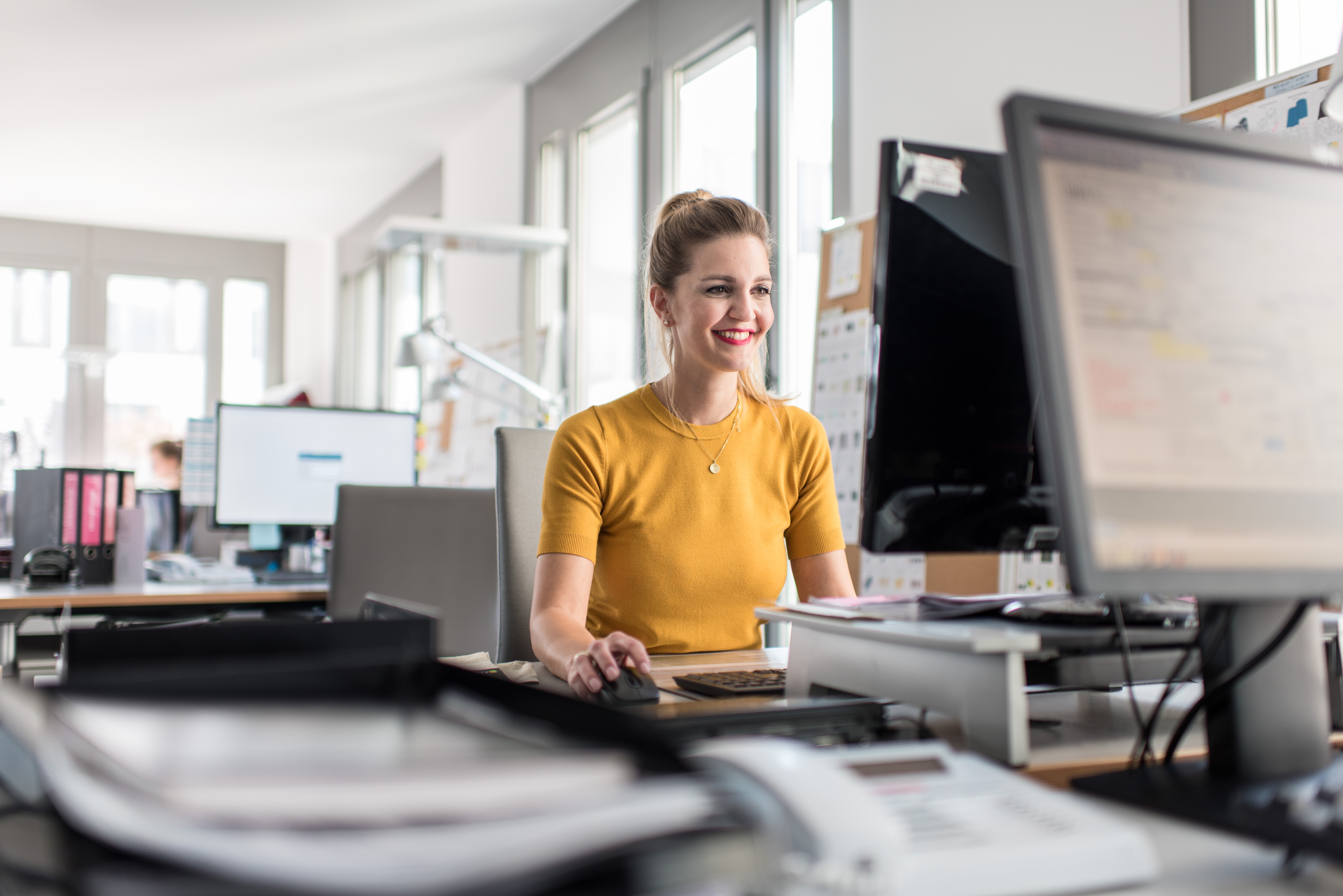 Eine Frau im gelben Shirt sitzt lächelnd am Schreibtisch und arbeitet am Computer.