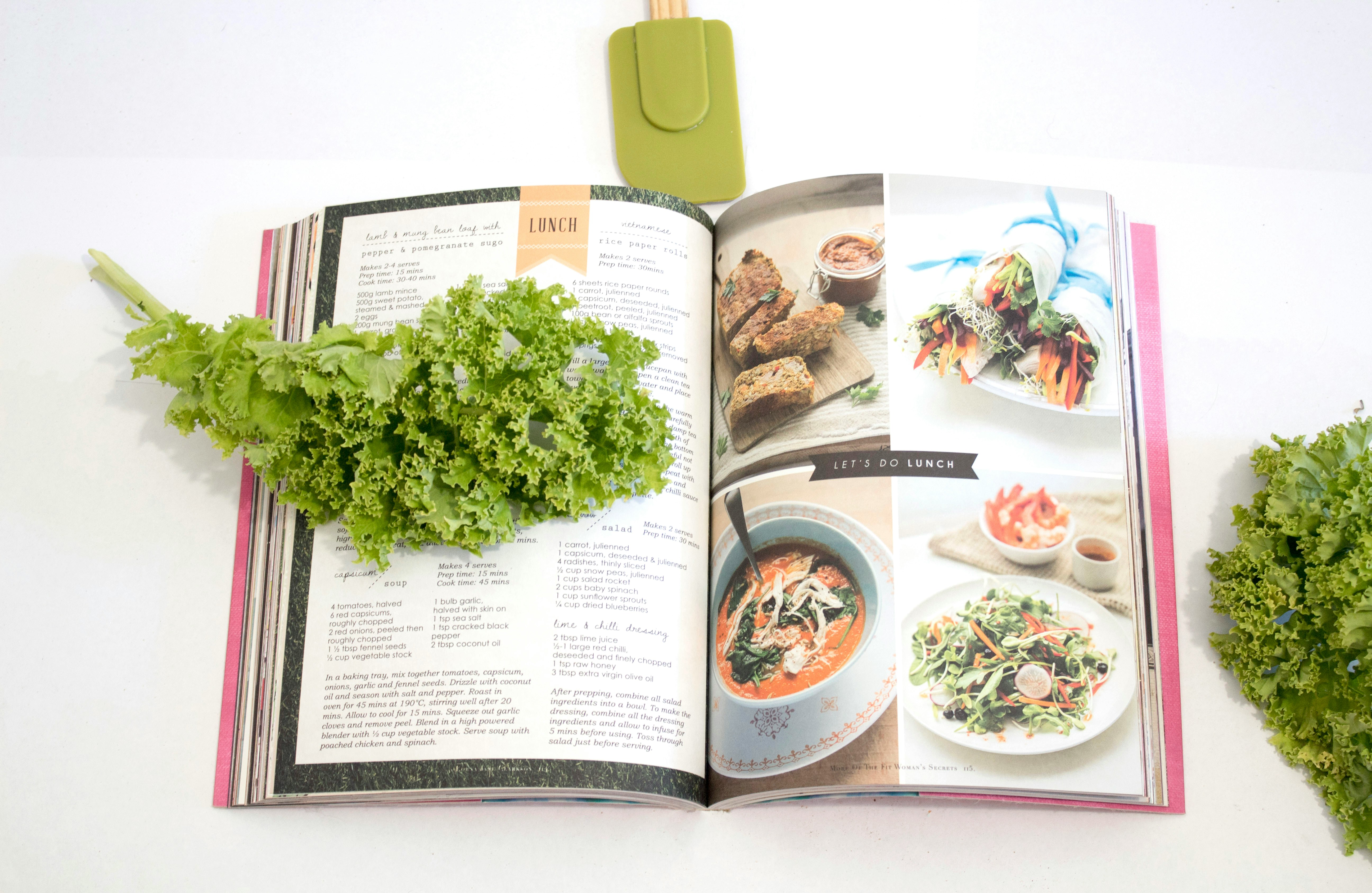 Ein Kochbuch liegt aufgeschlagen auf einem hellen Tisch, darauf liegt ein Salatblatt
