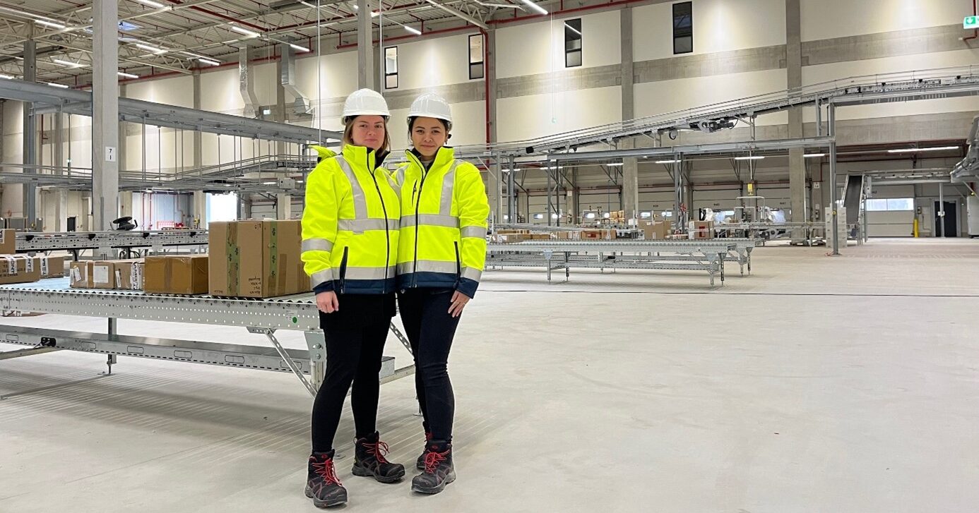 Die Logistikmitarbeiterinnen Franzi und Xuni stehen in Arbeitskleidung im Lager in Bedburg.